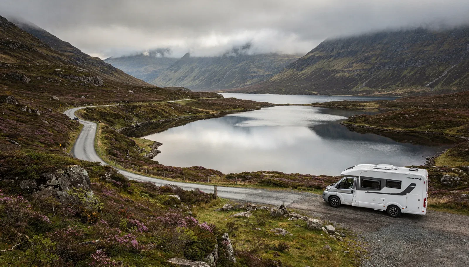 Route sinueuse à voie unique dans les Highlands écossais, camping-car garé face à un loch entouré de collines de bruyère